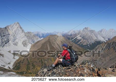 Male Hiker Sitting On Top Of Mountain Ridge Overlooking Valley And Mountains In The Distance With Blue Sky; Alberta Canada View Large Photo Image Stock Photo - Male Hiker Sitting On Top Of Mountain Ridge Overlooking Valley And Mountains In The Distance With Blue Sky; Alberta Canada. Fotosearch