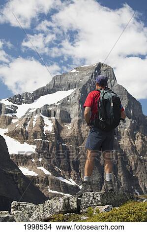 Male Hiker With A Backpack On Cliff Lookout Overlooking A Mountain Face With Snow; Lake Louise Alberta Canada View Large Photo Image Picture - Male Hiker With A Backpack On Cliff Lookout Overlooking A Mountain Face With Snow; Lake Louise Alberta Canada. Fotosearch
