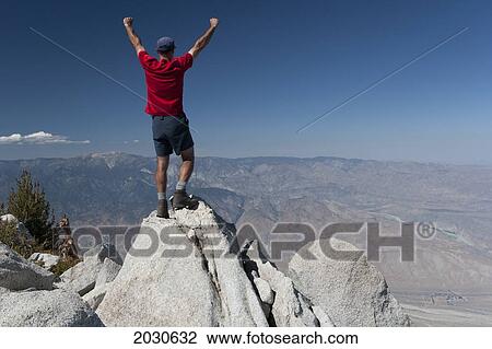 Stock Image - Male hiker with arms raised on top of desert mountain peak with mountain range and desert valley in distance and blue sky; palm springs california united states of america. Fotosearch