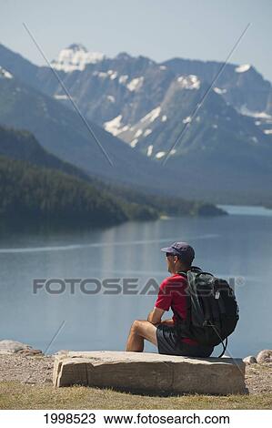 Male Hiker With Backpack Sitting On A Rock Looking Out Onto A Lake With Mountains And Blue Sky; Waterton Alberta Canada View Large Photo Image Stock Image - Male Hiker With Backpack Sitting On A Rock Looking Out Onto A Lake With Mountains And Blue Sky; Waterton Alberta Canada. Fotosearch