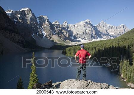 Man Standing On A Cliff Lookout Overlooking A Lake And Mountain Range With Blue Sky; Alberta Canada View Large Photo Image Stock Image - Man Standing On A Cliff Lookout Overlooking A Lake And Mountain Range With Blue Sky; Alberta Canada. Fotosearch