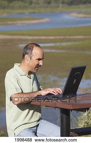 Stock Photo - Man Typing On A Laptop Sitting At A Picnic Table With A River Delta In The Background; Calgary Alberta Canada. Fotosearch
