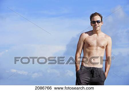 Stock Photograph - Portrait of a man wearing a swimsuit and sunglasses against a blue sky; punta cana la altagracia dominican republic. Fotosearch