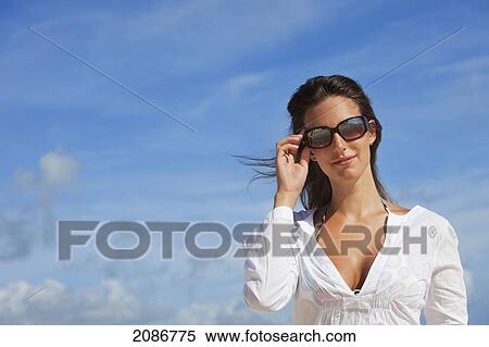 Stock Photography - Portrait of a woman wearing a white shirt and sunglasses against a blue sky; punta cana la altagracia dominican republic. Fotosearch