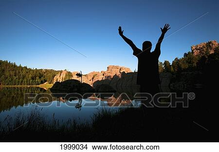 Picture - Silhouette Of A Young Man Standing At The Water's Edge Of Sylvan Lake With His Arm's Raised; Black Hills South Dakota United States Of America. Fotosearch