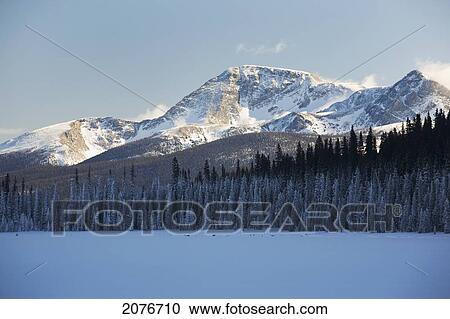 Sun lit snow covered mountain in the background with a snow covered lake and evergreen trees in the shade with blue sky; alberta canada View Large Photo Image Stock Image - Sun lit snow covered mountain in the background with a snow covered lake and evergreen trees in the shade with blue sky; alberta canada. Fotosearch