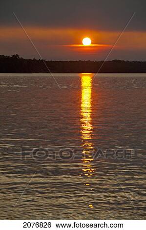Stock Photograph - Sunlight reflecting on a tranquil lake at sunset; lake of the woods ontario canada. Fotosearch
