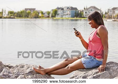 Stock Image - Teenage Girl Viewing Test Messages While Relaxing On The Shore Of A Residential Lake Area; Edmonton Alberta Canada. Fotosearch
