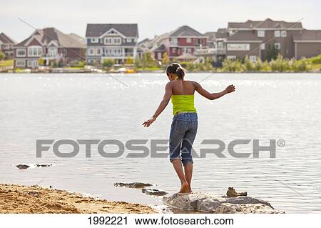 Stock Image - Teenage Girl Walking On The Rocks Along The Shore Of A Residential Lake; Edmonton Alberta Canada. Fotosearch