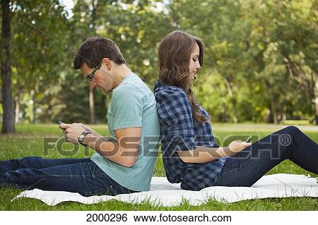 Stock Photograph - Young Couple Looking At Text Messages On Their Phones As They Sit Back To Back; Edmonton Alberta Canada. Fotosearch