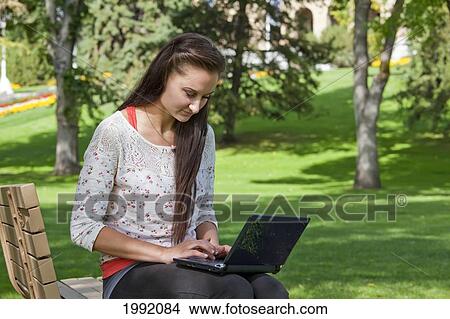 Picture - Young Woman Sitting On A Park Bench While Working On A Laptop; Edmonton Alberta Canada. Fotosearch