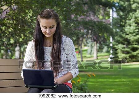 Stock Image - Young Woman Sitting On A Park Bench While Working On A Laptop; Edmonton Alberta Canada. Fotosearch