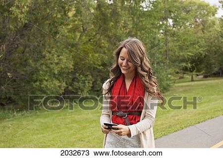 Stock Image - Young Woman Walking In A Park And Texting; Edmonton Alberta Canada. Fotosearch