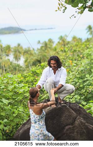 Stock Photography - A man helps a woman up onto a rock with a view of the ocean at bora bora nui resort and spa; bora bora island society islands french polynesia south pacific. Fotosearch