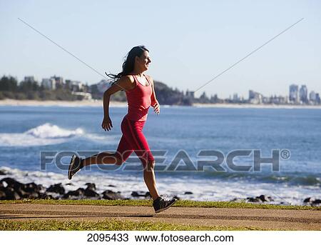 Stock Image - A woman runs on a path along the water; gold coast queensland australia. Fotosearch