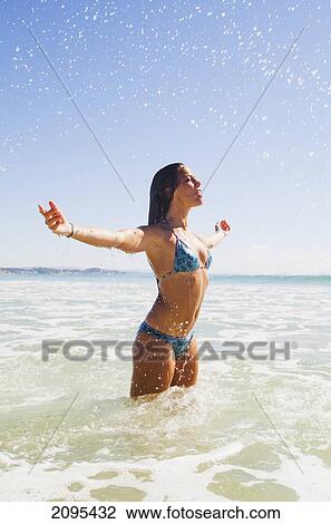 Stock Image - A young woman in a bikini splashes water as she stands in the ocean; gold coast queensland australia. Fotosearch