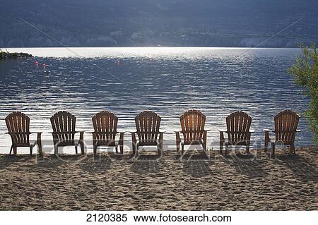 Stock Photography - Beach chairs lined up on the shoreline of a sun reflective lake with shadows of the chairs on the sand; summerland british columbia canada. Fotosearch