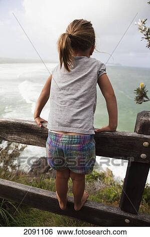Stock Photograph - Girl standing on the rail of a wooden fence looking out at the ocean; broken heads, new south wales, australia. Fotosearch