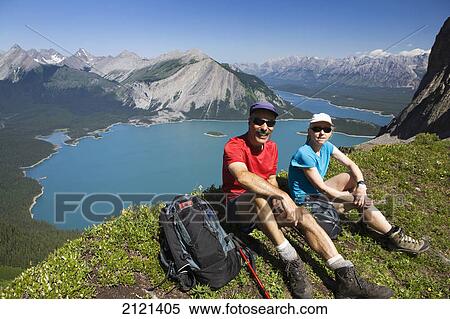 Stock Photography - Male and female hiker sitting on top of a mountain ridge overlooking emerald lakes and mountain ranges with blue sky; kananaskis provincial park alberta canada. Fotosearch