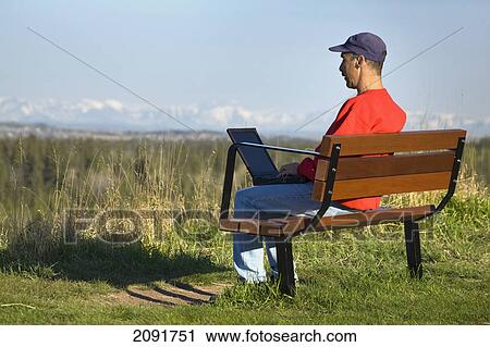 Man sitting on a bench in a park working on a laptop with snow covered mountains and blue sky in the background; calgary, alberta, canada View Large Photo Image Stock Image - Man sitting on a bench in a park working on a laptop with snow covered mountains and blue sky in the background; calgary, alberta, canada. Fotosearch