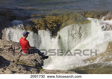 Stock Image - Man sitting on the edge of a cliff looking down on waterfalls at kananaskis provincial park; alberta, canada. Fotosearch