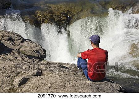 Stock Image - Man sitting on the edge of a cliff looking down on waterfalls in kananaskis provincial park; alberta, canada. Fotosearch