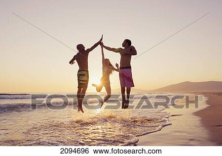 Stock Photograph - Three people leaping in the air to clap hands together on a beach at sunset; tarifa, cadiz, andalusia, spain. Fotosearch