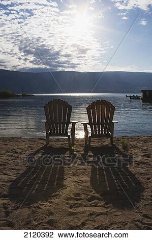 Stock Image - Two beach chairs on a beach with the sun burst out of the clouds and long shadows of the chairs on the sand; summerland british columbia canada. Fotosearch