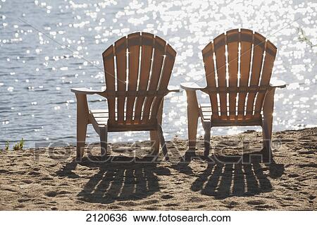 Stock Photograph - Two beach chairs on a sandy beach at the shoreline with sunlight reflecting off the lake and shadows of the chairs on the sand; summerland british columbia canada. Fotosearch