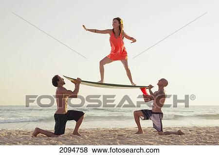 Two men hold a surfboard as a woman stands on it in a surfer pose; tarifa, cadiz, andalusia, spain View Large Photo Image Stock Photo - Two men hold a surfboard as a woman stands on it in a surfer pose; tarifa, cadiz, andalusia, spain. Fotosearch