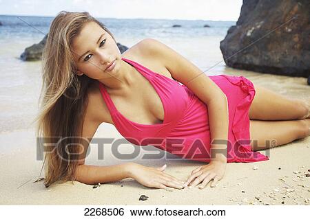 Stock Photograph - A young woman in a red swimsuit cover-up poses while laying in the sand on anini beach; kauai hawaii united states of america. Fotosearch