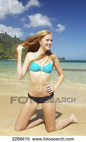 Stock Photography - A young woman kneeling in the sand by the ocean along the coast; kauai hawaii united states of america. Fotosearch