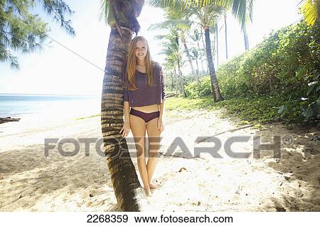 Stock Photo - A young woman poses beside a palm tree on a beach; kauai hawaii united states of america. Fotosearch