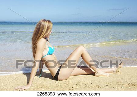 Stock Image - A young woman sitting in the sand along the ocean at the coast; kauai hawaii united states of america. Fotosearch