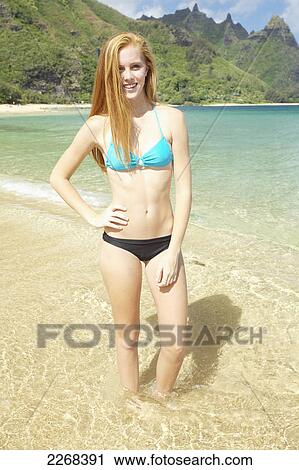 Stock Image - A young woman standing in the shallow water of the ocean along the coast; kauai hawaii united states of america. Fotosearch