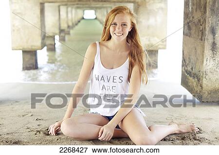 Stock Photo - A young woman with long red hair sits in the sand under a bridge at the water's edge; kauai hawaii united states of america. Fotosearch