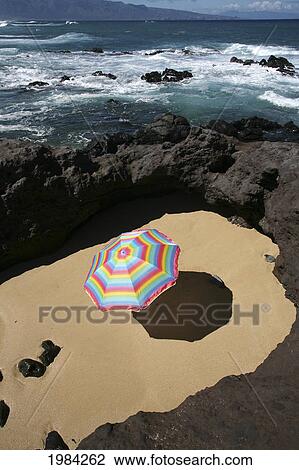 Stock Image - Brightly Colored Beach Umbrella In A Sandy Circle Surrounded By Lava Rock Next To The Ocean.. Fotosearch