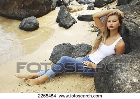 Picture - Portrait of a young woman sitting on the sand against a rock at the water's edge; kauai hawaii united states of america. Fotosearch