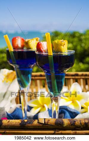 Stock Image - Two Tropical Drinks Garnished With Flowers And Fruit In An Outdoor Setting.. Fotosearch