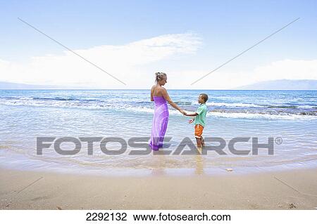Stock Image - A mother and son hold hands while standing in the shallow water at the ocean; Hawaii united states of america. Fotosearch
