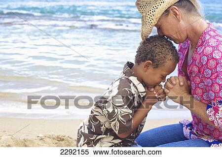 Stock Photography - A mother and son pray together on the beach at the water's edge; Hawaii united states of america. Fotosearch