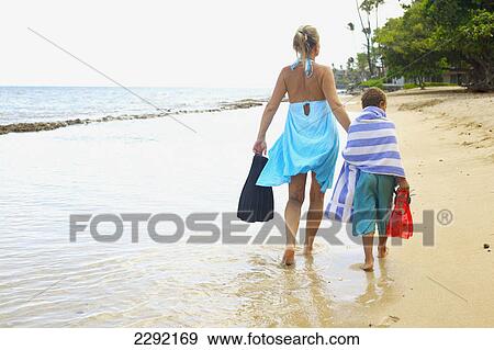 Stock Photo - A mother and son walking along the beach in swimsuits holding flippers; Hawaii united states of america. Fotosearch