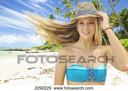 Stock Photo - A teenage girl on the beach in a two piece bathing suit and hat with her long hair blowing in the wind; Maui hawaii united states of america. Fotosearch