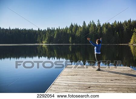 Picture - A woman greets the morning sun on a dock on lizard lake; Vancouver island british columbia canada. Fotosearch