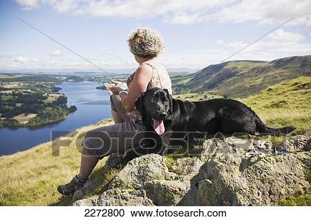 Stock Image - A woman sits with her dog on a rock overlooking a large lake and landscape; Hallin fell ullswater lake district england. Fotosearch