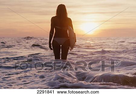 Stock Image - A woman standing in the ocean holding a surfboard at sunset; Canos de meca cadiz andalusia spain. Fotosearch