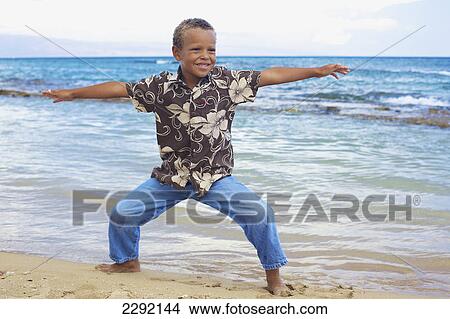Picture - A young boy does a yoga pose in the sand at the water's edge; Hawaii united states of america. Fotosearch