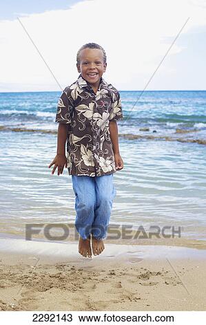 Stock Image - A young boy jumps with joy on the sand at the water's edge; Hawaii united states of america. Fotosearch