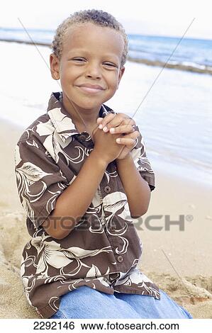 Stock Photograph - A young boy kneels with clasped hands in the sand at the water's edge; Hawaii united states of america. Fotosearch
