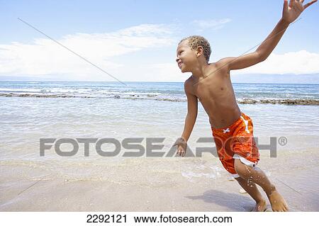 Stock Image - A young boy runs and plays in the shallow water of the ocean; Hawaii united states of america. Fotosearch